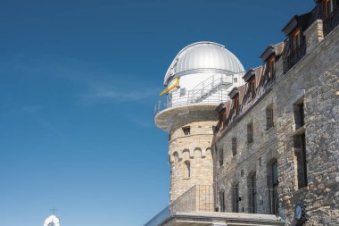 Gornergrat Rasathanesi, İsviçre 'nin Zermatt şehrinde yüksek irtifada yer alan bir tepede yer almaktadır ve arka planda bir taş cephe, modern kubbe ve bir şapel bulunmaktadır..
