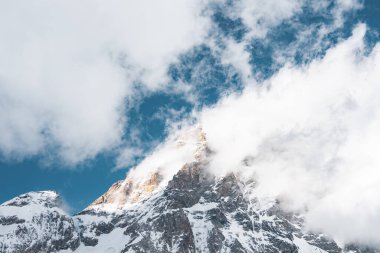 İsviçre 'nin Zermatt kenti yakınlarında karla kaplı bir dağ zirvesi, kısmen bulutlarla gizlenmiş. Pürüzlü yüz hatları ve güneş ışığı dramatik bir alp manzarası yaratır.