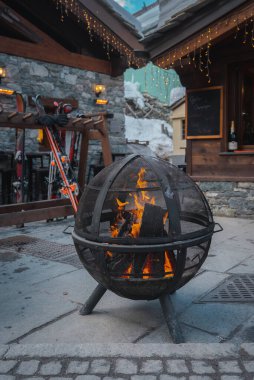 Yanan odunları olan metal bir ateş çukuru, ahşap bir kayak rafı ve İsviçre 'nin Zermatt şehrinde karlı bir dağda sicim ışıkları olan taştan bir bina..