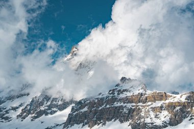 Simgesel Matterhorn dağı sisli bulutların arasından yükselir, Zermatt 'taki parlak mavi gökyüzünün altında ön planda kayalık yamaçlar vardır..