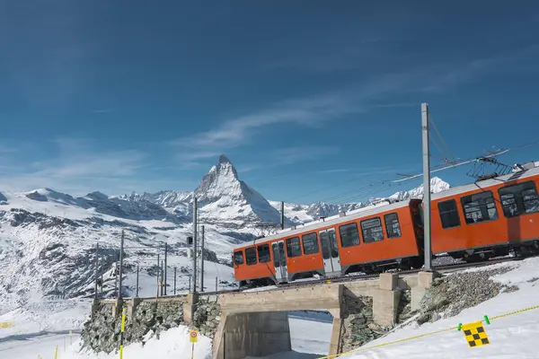 Karlı Zermatt, İsviçre 'de küçük bir köprüden geçen kırmızı bir tren, Matterhorn dağı ve açık mavi gökyüzü çarpıcı bir alp manzarası yaratıyor..