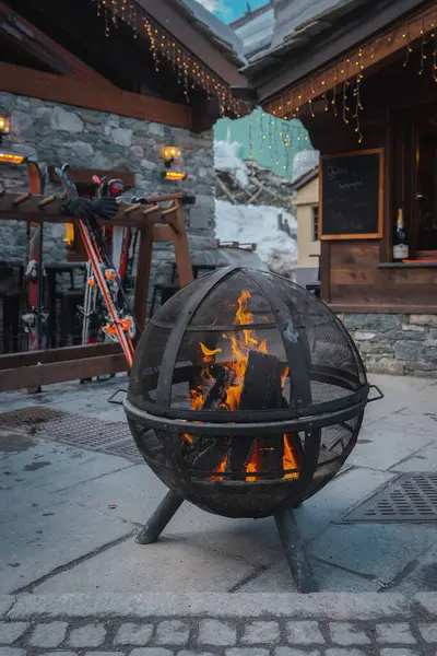 Yanan odunları olan metal bir ateş çukuru, ahşap bir kayak rafı ve İsviçre 'nin Zermatt şehrinde karlı bir dağda sicim ışıkları olan taştan bir bina..