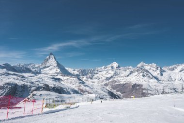 İsviçre 'deki Zermatt kayak merkezi, açık mavi gökyüzünün altında Matterhorn, kar kaplı yamaçlar, kayak asansörleri, kayakçılar ve alp zirvelerine sahiptir..