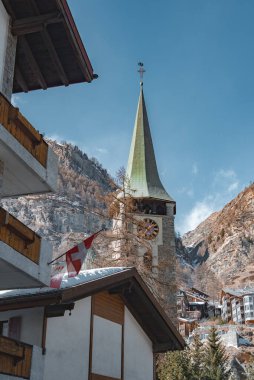 St. Mauritius Kilisesi 'nin yeşil kule ve saat çehresi, arka planda kar tozlu dağlar ve açık mavi gökyüzü ile dağlık binaların arasında göze çarpıyor..