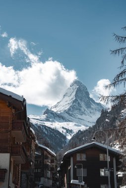 İsviçre, Zermatt 'taki Matterhorn dağınık bulutlu mavi gökyüzüne karşı yükseliyor. Tahta kadehler ve çıplak ağaçlar kışın dağlık sahnenin çerçevesini oluşturuyor..