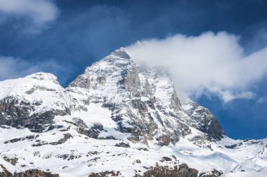 İsviçre, Zermatt yakınlarındaki ikonik Matterhorn dağı, kar kaplı yamaçlar, engebeli kaya oluşumları ve mavi gökyüzünün altında ince bir bulut tabakası..