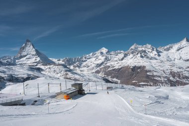 Kar yamaçları, kayak asansörleri ve Zermatt kayak merkezi sahnesine hakim, arka planda açık gökyüzü ve alp zirveleri var..