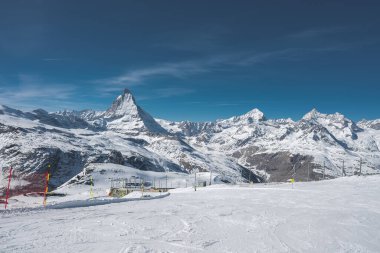 İsviçre 'deki Zermatt kayak merkezinin manzarasında açık mavi gökyüzü altında Matterhorn, kayak asansörleri, karla kaplı yamaçlar ve engebeli alp zirveleri görülüyor..