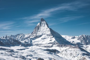 İkonik Matterhorn dağı, Zermatt yakınlarındaki İsviçre Alplerinde kar dolu tepe ve vadilerle çevrili açık mavi gökyüzüne karşı keskin bir şekilde yükselir..