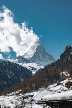 İsviçre 'nin Zermatt kenti yakınlarındaki ikonik Matterhorn dağı, ağaçlarla çevrili, kayalık arazi ve ahşap bir yapı ile keskin bir şekilde yükselir..