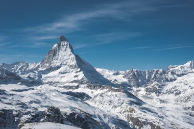 İkonik Matterhorn dağı, açık mavi gökyüzü altında kar, buzullar ve engebeli dağlarla çevrili piramit şekliyle keskin bir şekilde yükselir..