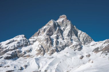İsviçre, Zermatt yakınlarındaki ikonik Matterhorn dağı, pürüzlü, karlı yamaçlarla çevrili açık mavi bir gökyüzüne karşı karla kaplı zirvelerle yükselir..