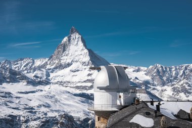 Matterhorn Piramidi, açık mavi gökyüzünün altında yükselen tepe gibi karlı dağlık arazide beyaz kubbeli bir gözlemevi üzerine kurulur..