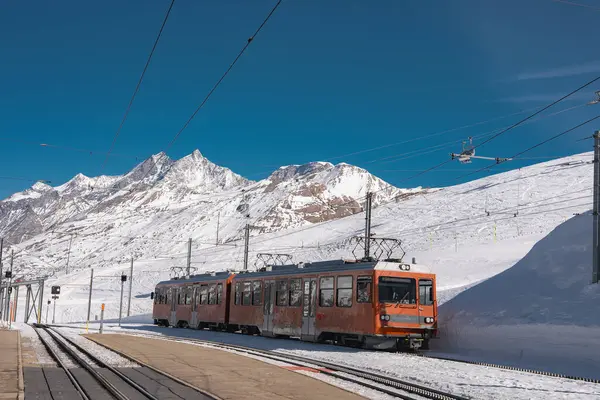 İsviçre, Zermatt 'ta parlak turuncu bir tren duruyor. Matterhorn aralığı, kayak asansörleri ve arka planda açık mavi gökyüzü var..