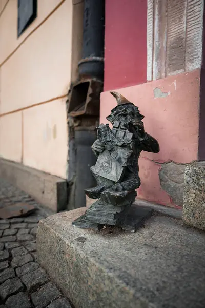 Polonya 'nın Wroclaw şehrinde kaldırım taşı caddesi boyunca taştan bir kaide üzerinde küçük bir bronz cüce heykeli. Arka planda bej ve kırmızı renkli bir bina duvarı var..