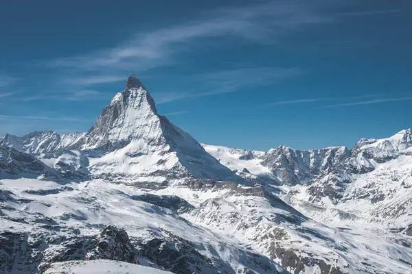 İkonik Matterhorn dağı, açık mavi gökyüzü altında kar, buzullar ve engebeli dağlarla çevrili piramit şekliyle keskin bir şekilde yükselir..