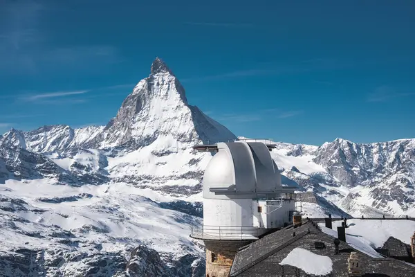 Matterhorn Piramidi, açık mavi gökyüzünün altında yükselen tepe gibi karlı dağlık arazide beyaz kubbeli bir gözlemevi üzerine kurulur..