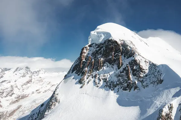 Kar, dağların zirvesini engebeli kaya yüzeyi ve açık mavi gökyüzünün altında Zermatt yakınlarındaki İsviçre Alplerinde dağılmış bulutlarla kapladı..
