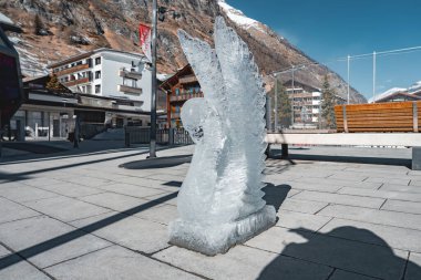 İsviçre 'nin Zermatt kentindeki kaldırımlı bir meydanda kanatlı bir meleğin detaylı buzdan heykeli. Dağlık binalar ve karla kaplı dağlarla çevrili..
