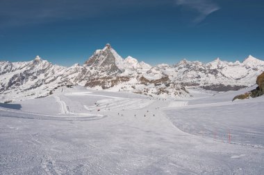 İsviçre 'deki Zermatt kayak merkezinin manzarası. Matterhorn, işaretli yamaçlarda kayakçılar, karla kaplı zirveler ve açık mavi bir gökyüzü..