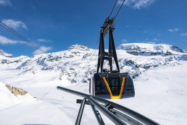 Pürüzsüz, siyah ve sarı bir teleferik İsviçre 'nin Zermatt kentindeki karla kaplı zirvelerde, açık mavi gökyüzü ve engebeli dağlık arazisiyle yükseliyor..