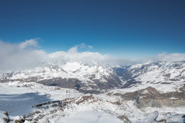İsviçre, Zermatt yakınlarında açık mavi bulutlu bir gökyüzünün altında kar, dağlık tepeleri ve buzulları kapladı. Engebeli arazi ve vadiler dramatik kontrastlar ekler.