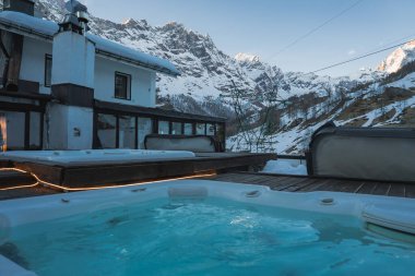 Karla çevrili ahşap bir güvertede lüks bir jakuzi, dağ tepeleri, dağ evi tarzı bir bina, kayak kaldırma kabloları ve Zermatt 'taki engebeli dağ yamaçları..