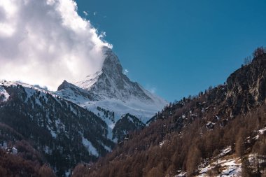 İsviçre, Zermatt yakınlarındaki ikonik Matterhorn, kar kaplı yamaçlar, yoğun ormanlar, engebeli uçurumlar ve berrak mavi gökyüzünün altındaki bulutlar..