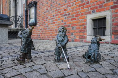 Polonya, Wroclaw 'da bir kaldırım taşı caddesinde çeşitli pozlarda üç bronz cüce heykeli, arka planda kırmızı tuğla bir duvar ve parmaklıklı pencere var..