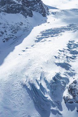İsviçre, Zermatt yakınlarındaki bir buzulda buzlu yarıklar, engebeli tepeler, kayalık uçurumlar ve dağlık araziye gölge düşüren güneş ışığı yer alıyor..