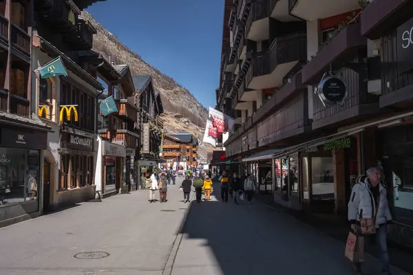 İsviçre, Zermatt 'ta hareketli bir cadde, dağ evi tarzı binalar, bir McDonalds, dükkanlar, yayalar ve gün ışığında karla kaplı dağlar..