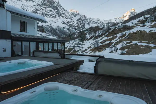 Tahta bir güvertede jakuziler Zermatt 'ta karla kaplı dağlara bakar. Beyaz bir bina ve kayak kaldırma kabloları kayak merkezi ayarlarını vurgular.