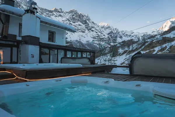 Karla çevrili ahşap bir güvertede lüks bir jakuzi, dağ tepeleri, dağ evi tarzı bir bina, kayak kaldırma kabloları ve Zermatt 'taki engebeli dağ yamaçları..