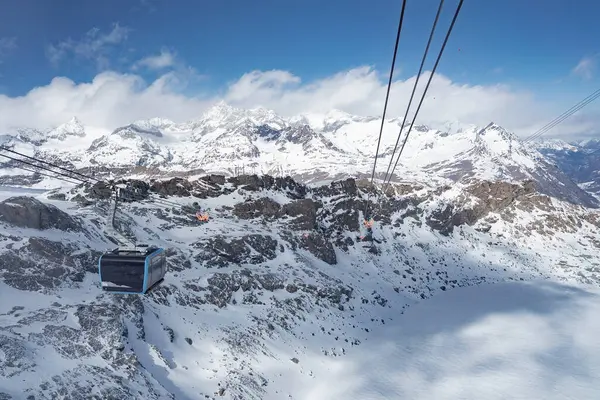 Zermatt kayak merkezi bölgesinde, karlı zirveler, buzullar ve parlak mavi gökyüzünün altındaki engebeli araziyle çevrili bir teleferik havada asılı kaldı..