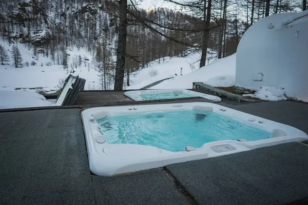 Açık havada bir jakuzi ve küçük bir havuz ahşap bir güvertede karla kaplı ağaçlar ve arka planda bir kış otelinde bir yamaçla çevrili..
