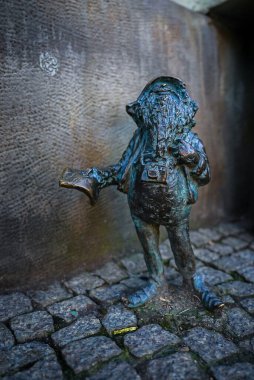 Polonya 'nın Wroclaw kentindeki dokulu bir duvara karşı elinde bir kitap tutan ve kamera takan küçük bronz bir cüce heykeli kaldırım taşları üzerinde duruyor..