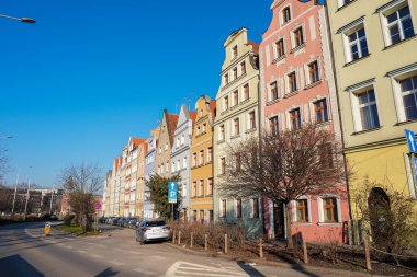 Wroclaw, Polonya 'da açık mavi gökyüzünün altında bir sıra pastel renkli, gaçalı bina. Park etmiş arabaların ve yapraksız ağaçların olduğu sessiz bir cadde görülebilir..