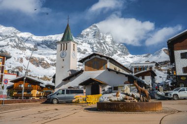Alplerdeki Breuil Cervinia manzarası, yeşil çan kulesine sahip beyaz bir kilise, dağlık binalar, dolambaçlı bir heykel ve Matterhorn Dağı..