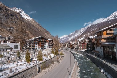 Zermatt, İsviçre manzarası, bir nehir, dağlık binalar, karla kaplı bir mezarlık, ve berrak gökyüzünün altındaki dağlar.