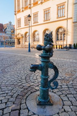 Polonya 'nın Wroclaw kentindeki bir kaldırım taşı meydanında, dekoratif bir su pompasının üzerinde duran bronz bir cüce heykeli, arka planda tarihi binalar bulunuyor..