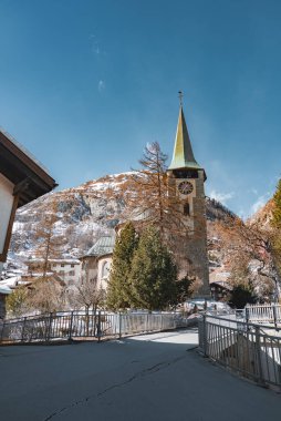 Yeşil kulesi ve saat yüzlü taş bir kilise engebeli dağlar ve açık mavi gökyüzü tarafından çerçevelenmiş kar tozlu ağaçların ve dağlık binaların arasında duruyor..