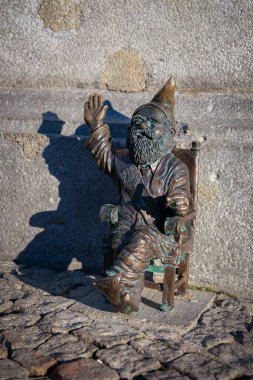 Küçük bronz bir cüce heykel bir sandalyede oturur ve tek eliyle el sallar. Polonya 'nın Wroclaw kentinde kaldırım taşlarının üzerine yerleştirilmiş ayrıntılı bir sakal ve şapka yer alıyor..