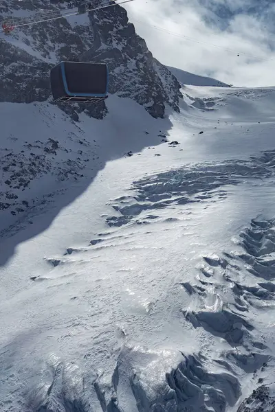 Modern bir teleferik gondolu, pürüzlü buz oluşumları, kayalık dağ yamaçları ve İsviçre 'nin Zermatt kentindeki dağınık bulutlarla karlı bir buzulun üzerinde seyahat eder..