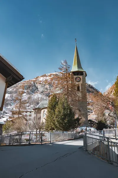 Yeşil kulesi ve saat yüzlü taş bir kilise engebeli dağlar ve açık mavi gökyüzü tarafından çerçevelenmiş kar tozlu ağaçların ve dağlık binaların arasında duruyor..