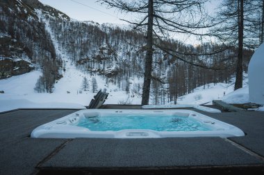 Tahta güvertede bir jakuzi, buhar yükseliyor, etrafı karla kaplı yamaçlar, seyrek ağaçlar ve İsviçre 'nin Zermatt kentindeki engebeli dağ arazileriyle çevrili..