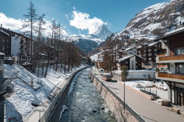 Zermatt, İsviçre manzarası, Matterhorn, temiz bir nehir, dağlık binalar, karlı bankalar, yapraksız ağaçlar, bir yol ve mavi gökyüzünün altında bir otobüs..