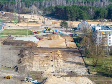 Riga, Letonya 'da inşaat alanı, ağır makineler, kum yığınları, elektrik direkleri, ormanlık alan ve arka planda bir yerleşim alanı gösteriyor..