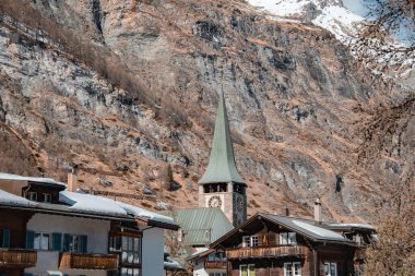 Yeşil kule ve saat yüzlü bir kilise manzarası, karla kaplı ahşap tebeşirle. Zermatt 'ın arka planında engebeli bir dağ yükseliyor.