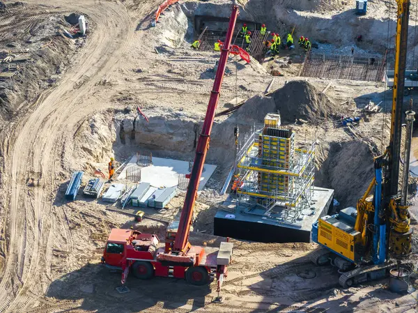 Riga, Letonya 'daki aktif inşaat alanında kırmızı bir vinç, sarı sondaj makinesi, işçiler, güçlendirilmiş çelik yapılar ve dağınık malzemeler görülüyor..