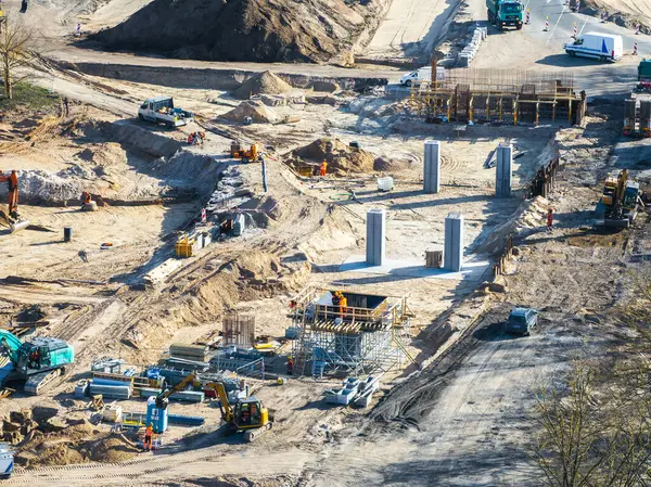 Riga, Letonya 'daki inşaat sahasında kazıcılar, kamyonlar, işçiler, beton sütunlar, iskeleler, toprak yığınları ve kısmen asfalt yollar görülüyor..